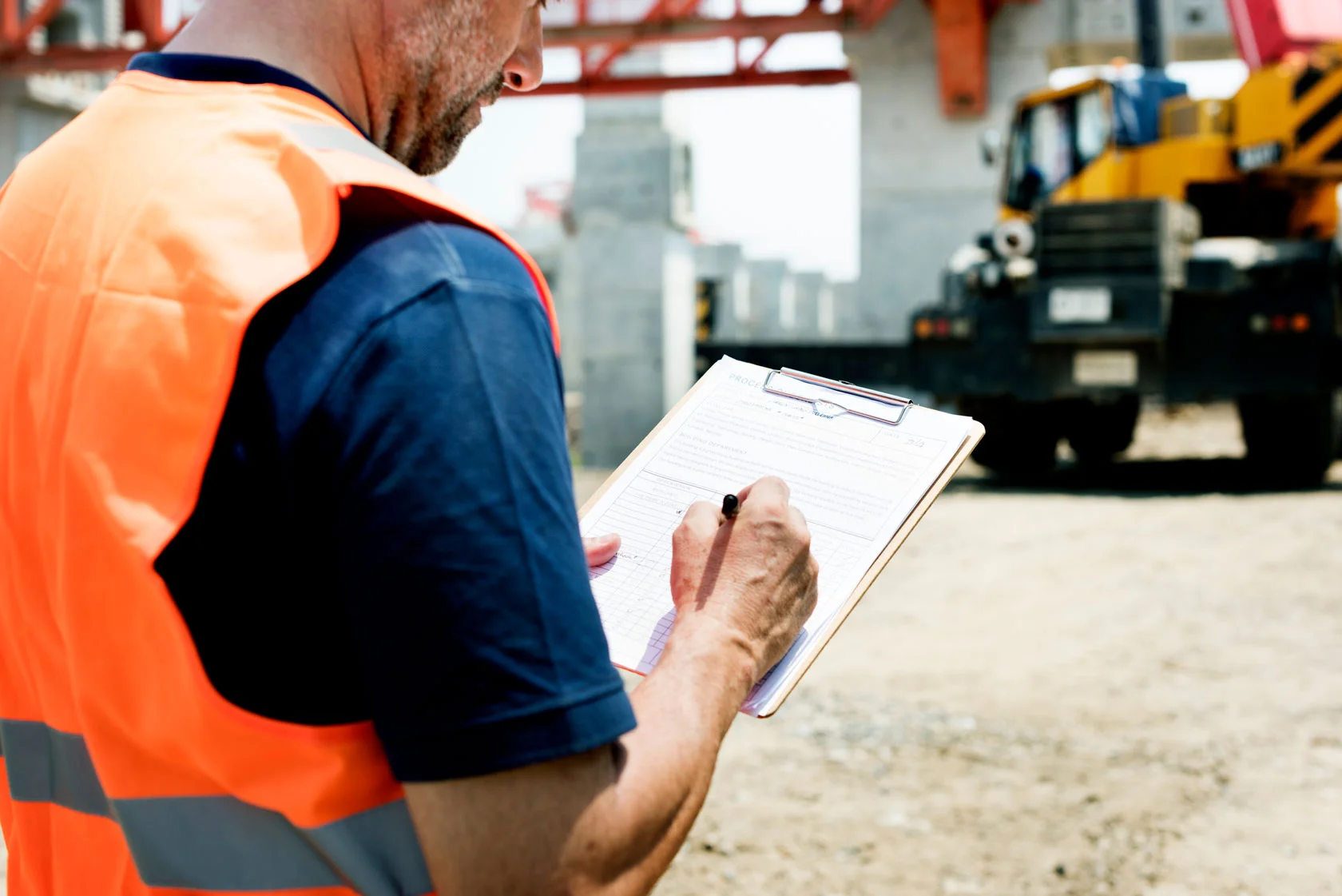 A construction worker wearing an orange safety vest writes on a clipboard at a building site, with machinery and unfinished structures visible in the background.