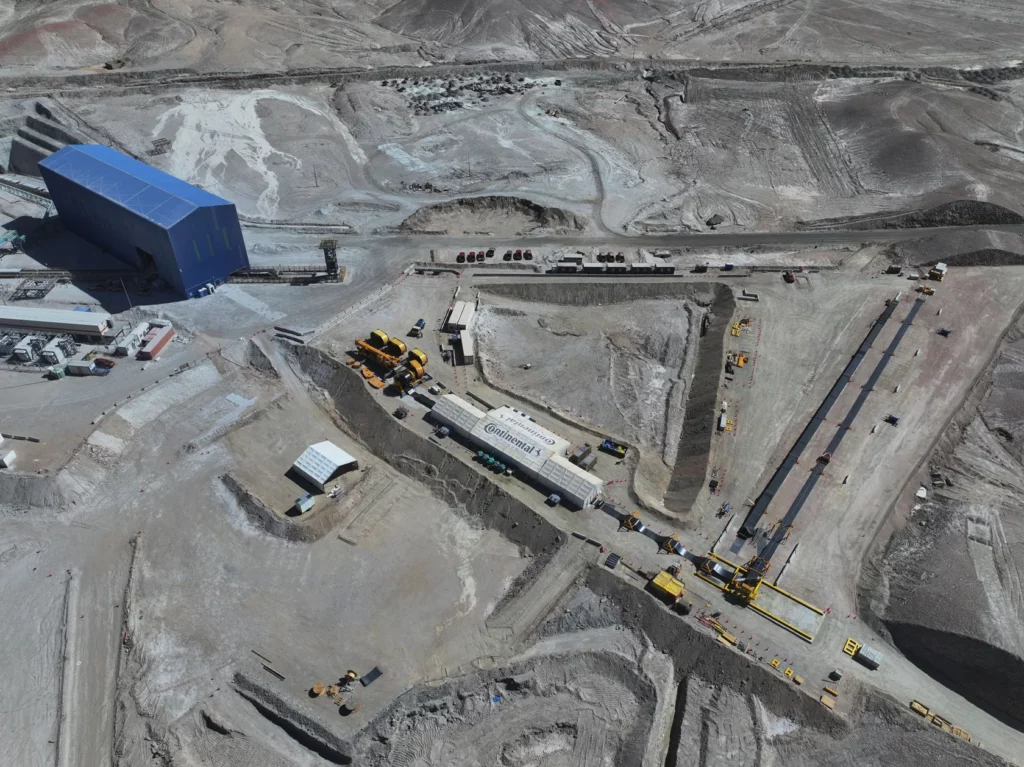 Aerial view of a large industrial mining site with a blue warehouse, heavy machinery, lorries, and excavated earth in a barren, rocky landscape. Workers and equipment are dispersed across the construction area.