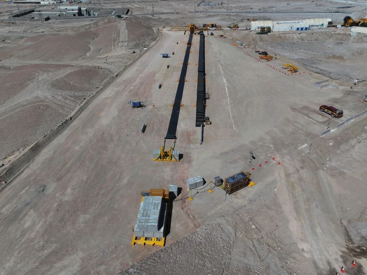 Aerial view of a construction site in a desert area with long steel beams laid out on the ground, machinery, vehicles, and equipment scattered around, and several buildings in the background.