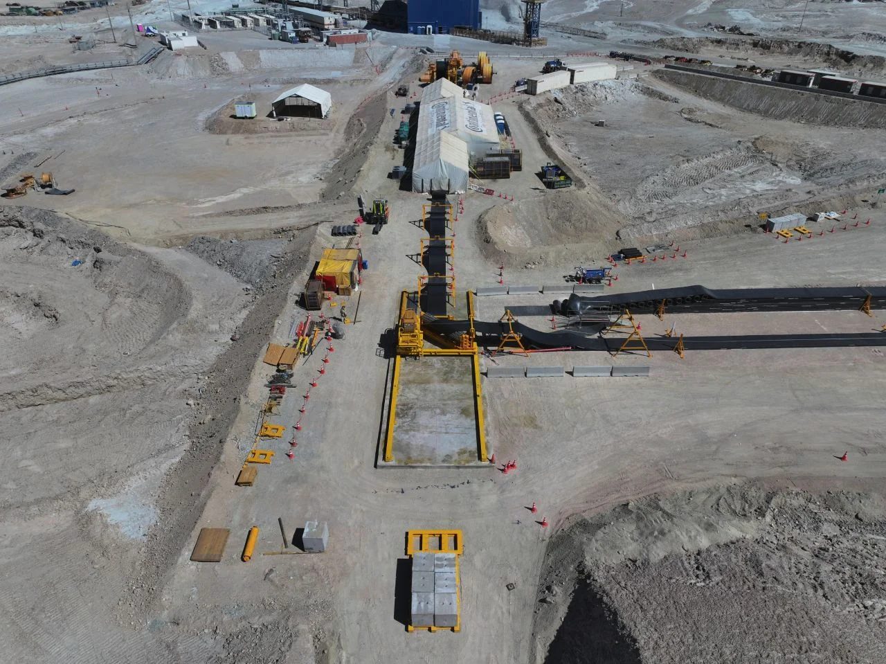 Aerial view of a large construction site in a dry, barren area with machinery, large marquees, equipment, and organised work zones. Dirt roads and construction materials are scattered throughout the site.