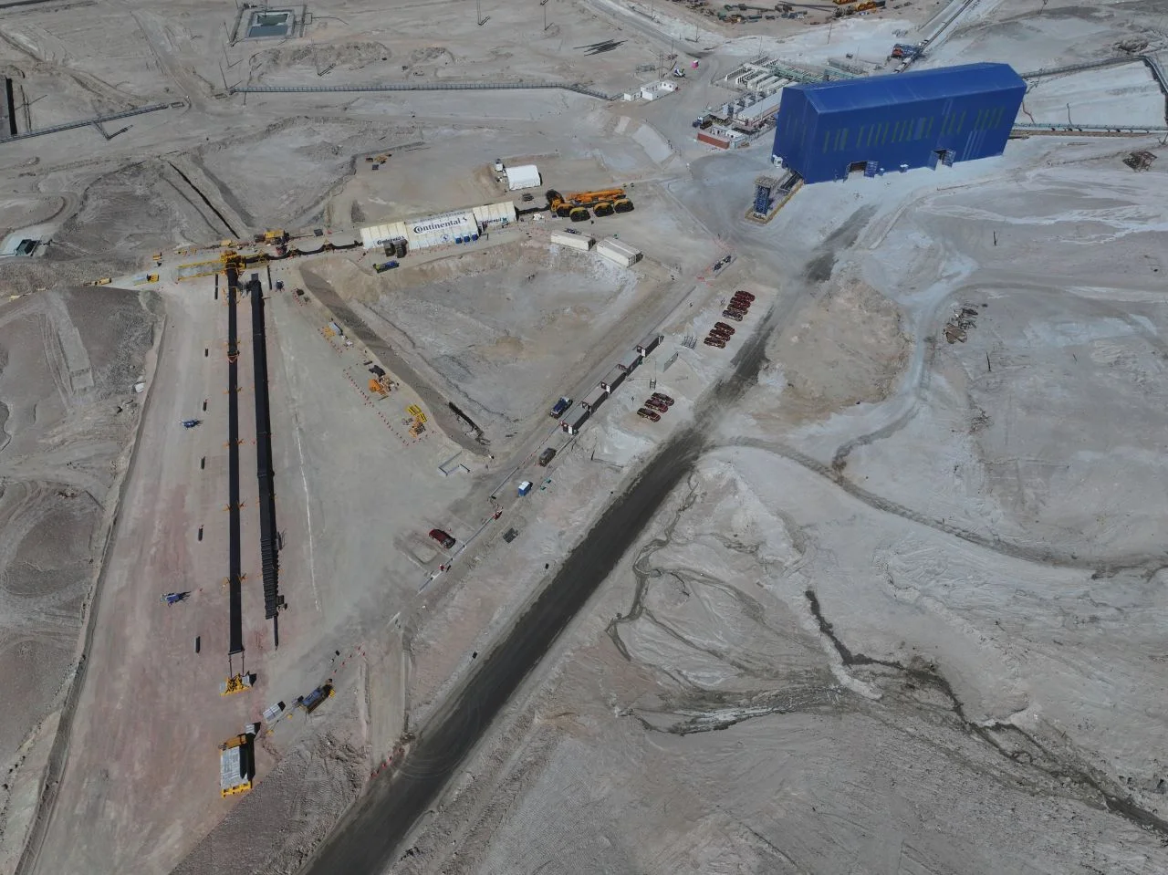 Aerial view of a large construction site in a desert area with a prominent blue building, multiple vehicles, scattered equipment, and dirt roads criss-crossing the barren, sandy landscape.