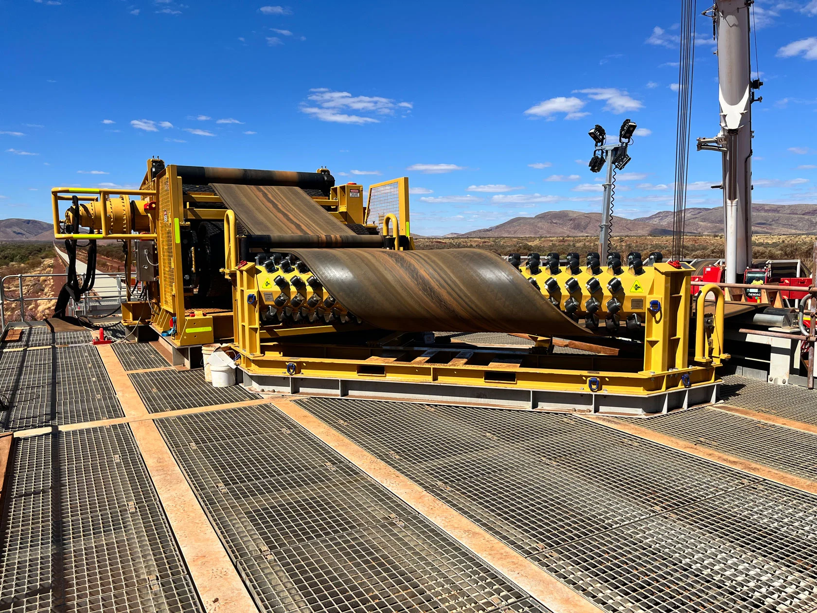 A large industrial conveyor belt machine with yellow metal framing is installed on a gritted platform outdoors, under a blue sky with scattered clouds and mountains in the background.
