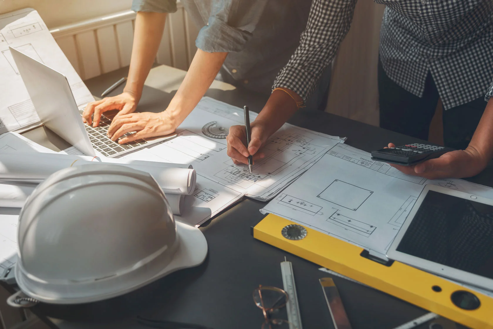 Two people work on architectural blueprints at a table cluttered with a hard hat, laptop, calculator, and measuring tools, suggesting a collaborative design or engineering project.