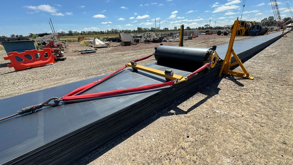 A long sheet of black conveyor belt material is unrolled at an outdoor industrial site, with machinery and red straps holding it in place on a gravel surface under a blue sky.
