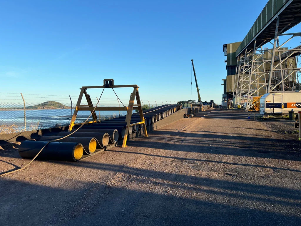 Long black pipes lie stacked beside a waterfront industrial site with fencing, construction vehicles, and a crane. A large metal structure runs parallel to the water under a clear blue sky.