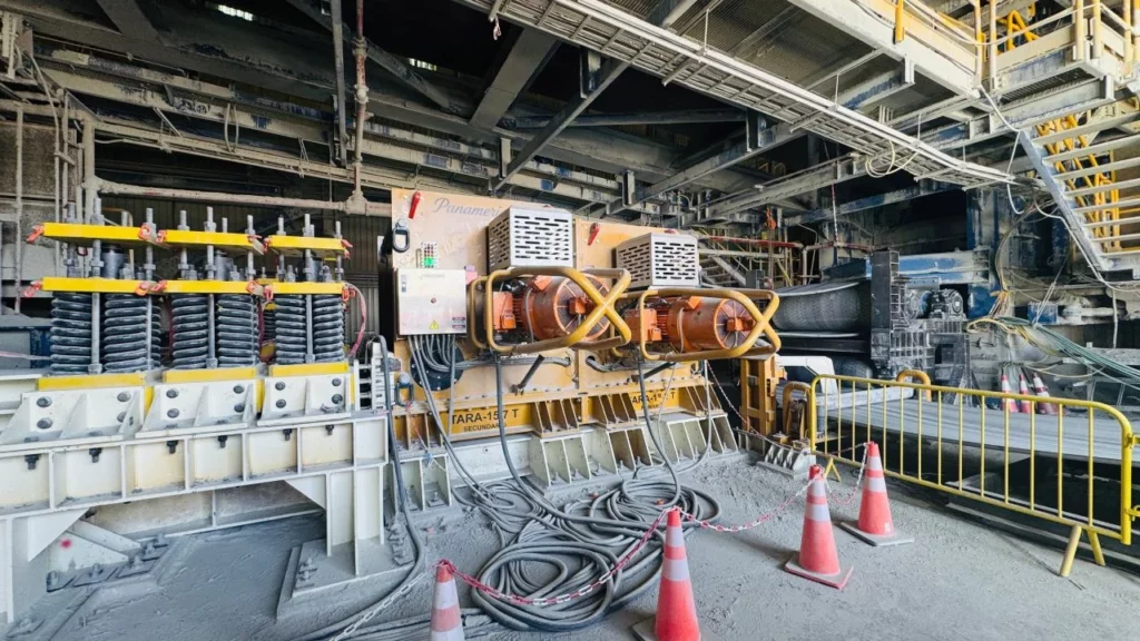Industrial interior with large machinery, cables on the floor, orange fans, traffic cones, and yellow safety barriers. Metal beams and platforms are visible above, and the area appears dusty and well-used.