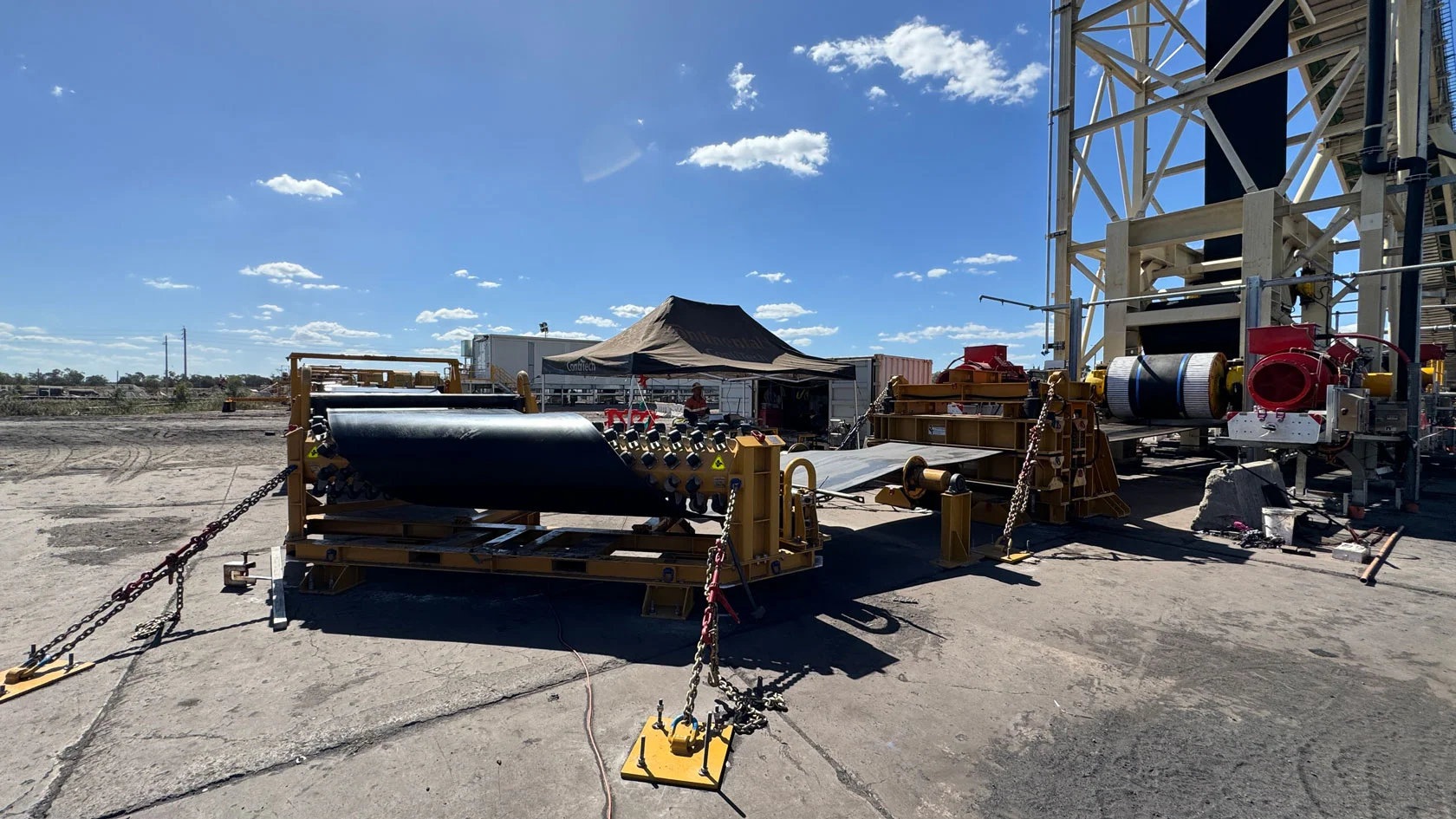 Outdoor industrial site with large machinery and conveyor belts, surrounded by chains and safety barriers. A marquee, fencing, and various equipment are visible under a bright blue sky with scattered clouds.