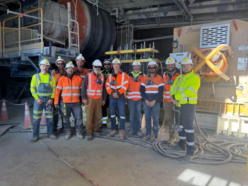 A group of workers in safety gear, including hard hats and high-visibility jackets, stand together at an industrial worksite with machinery and cables visible in the background.