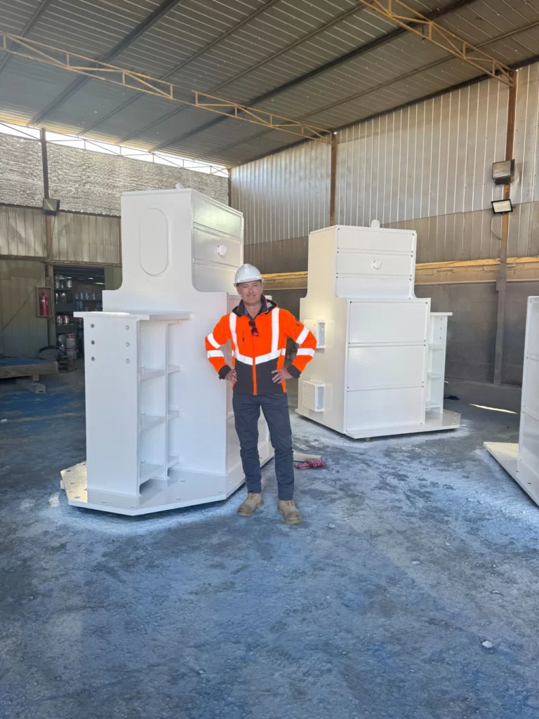 A person in a high-visibility orange jacket and white hard hat stands inside an industrial warehouse, surrounded by large, white-painted metal structures.