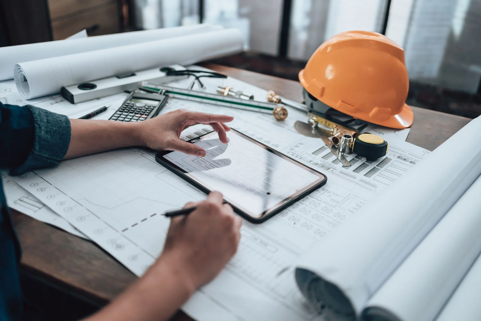 A person reviews architectural plans on a desk, using a tablet and making notes. The desk holds a yellow hard hat, ruler, calculator, tape measure, and other drawing tools.