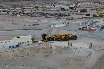 Two large yellow mining lorries are parked on a dusty construction site surrounded by equipment, containers, and industrial buildings in a barren, rocky landscape.