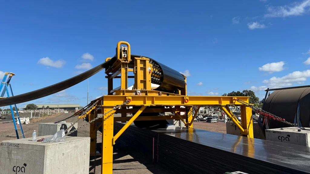 A large yellow industrial machine unwinds and processes a roll of black material outdoors under a blue sky, with buildings and equipment visible in the background.