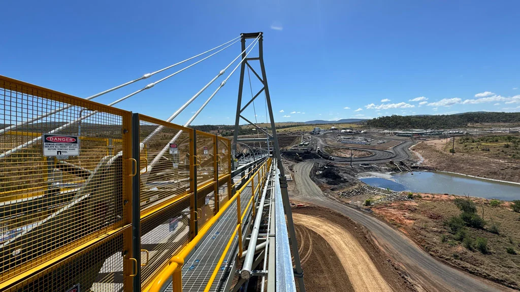 View from a high metal walkway with railings overlooking a large mining site, dirt tracks, ponds, and distant hills under a clear blue sky. Warning signs are visible on the yellow railing.
