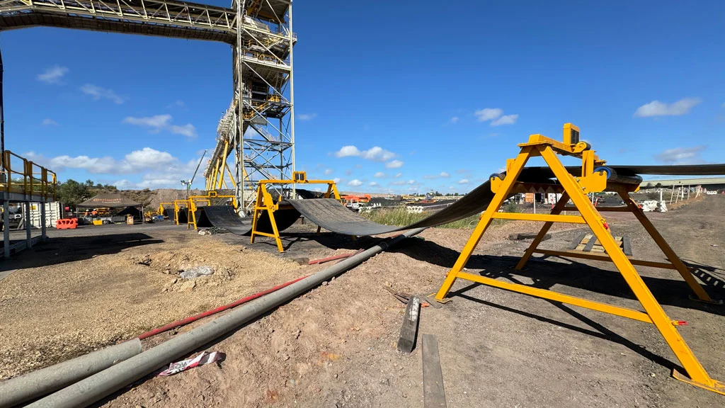 A damaged conveyor belt lies off its track at an outdoor industrial site under a blue sky, with yellow support structures and scattered construction materials visible.