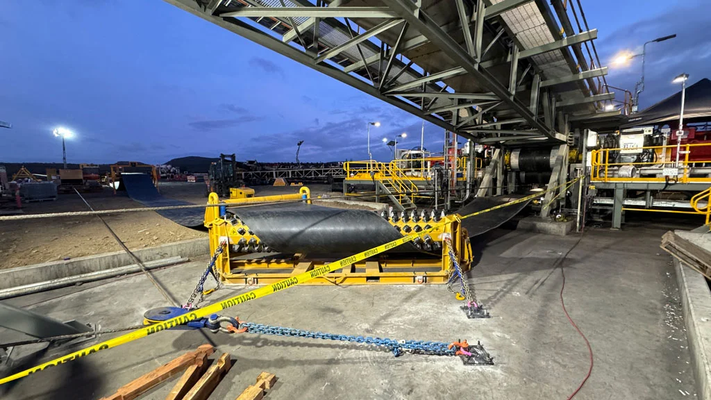 A construction site at dusk shows a large conveyor structure overhead, yellow warning tape and chains blocking access, and industrial equipment and materials arranged on the ground. Bright lights illuminate the work area.
