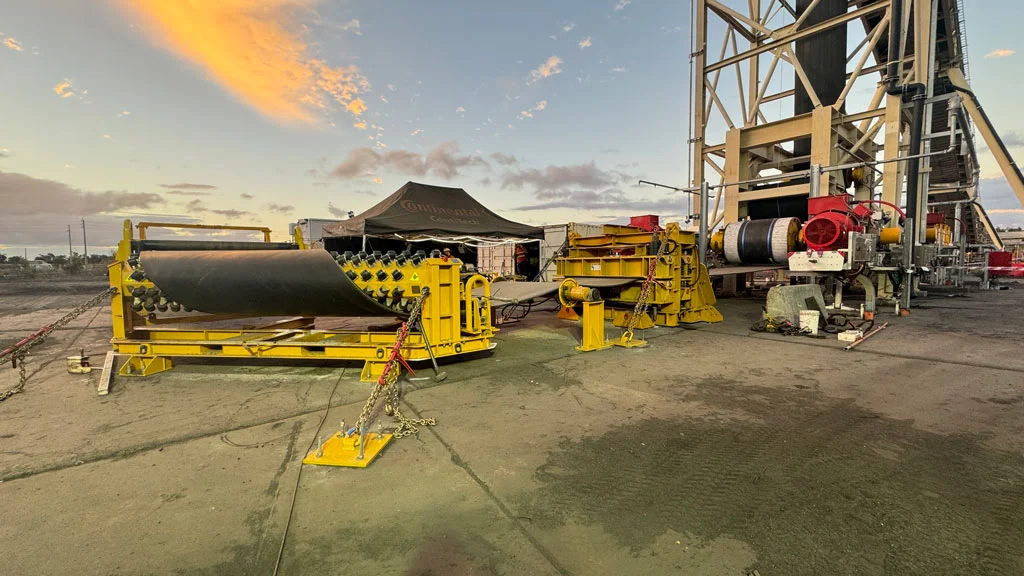 Industrial equipment, including a large black conveyor belt on yellow supports, set up outdoors near a tall metal structure at sunset; tents and more machinery are visible in the background.