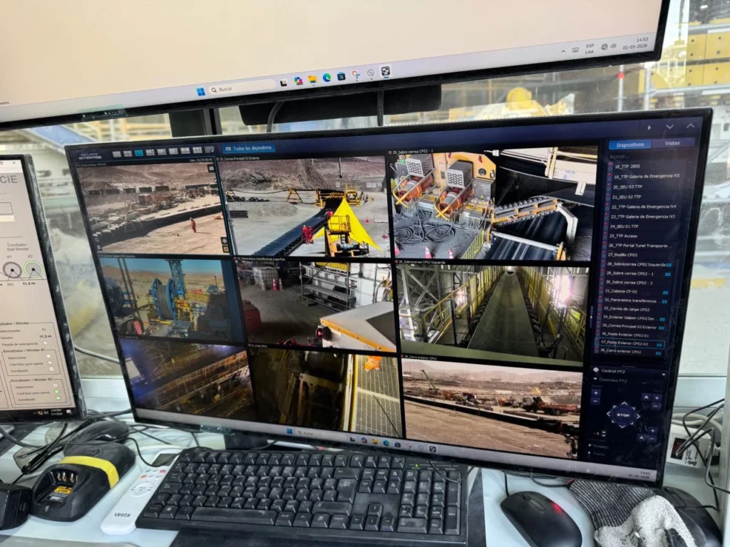 A computer monitor displays a grid of surveillance camera feeds showing various industrial and construction site areas, with a keyboard, mouse, and other office equipment in front of it.