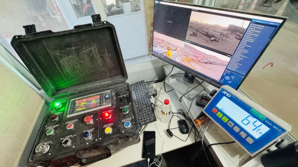 A control station with a keyboard, control panel with buttons and dials, a computer monitor showing CCTV footage of an industrial site, and an electronic scale displaying 64.1 kg.