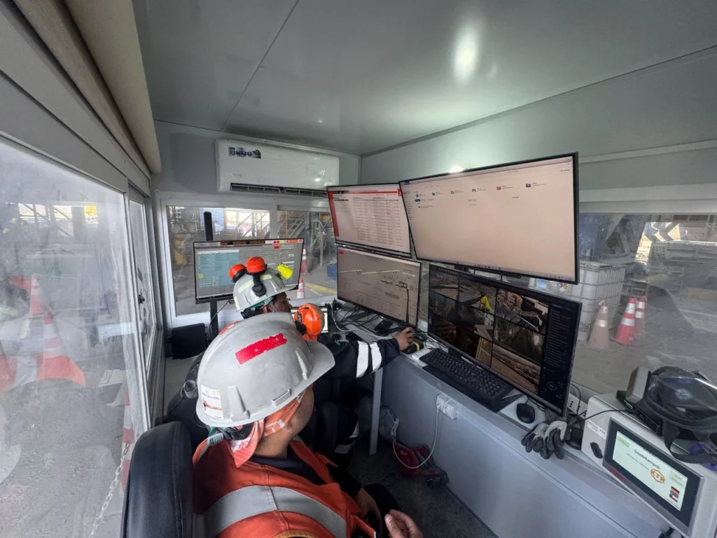 Two workers wearing safety gear and helmets operate multiple computer monitors in a control room, overseeing industrial operations through CCTV cameras and data screens. Traffic cones and equipment are visible outside the window.