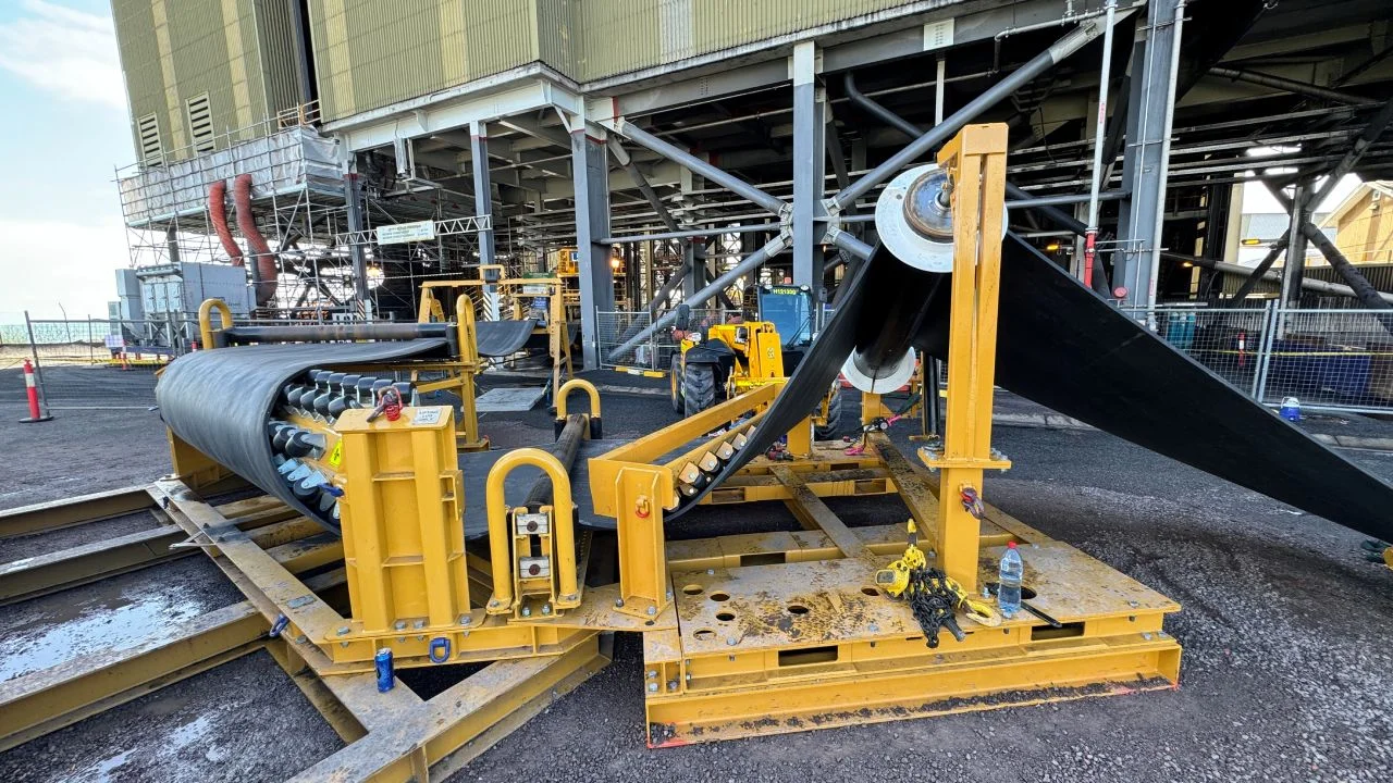 Large yellow machinery is set up outdoors, feeding rolls of black conveyor belt material under a raised industrial structure, with equipment and construction materials scattered around the site.
