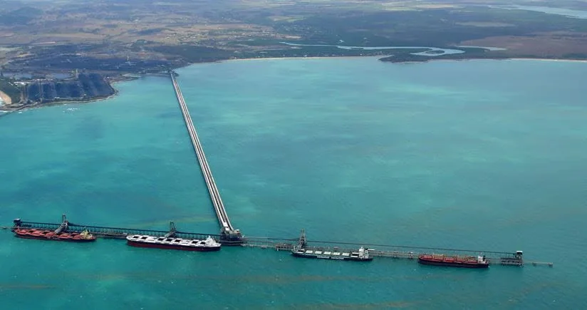 Aerial view of a long pier extending into turquoise water, with several large cargo ships berthed at the end, and coastline visible in the background.