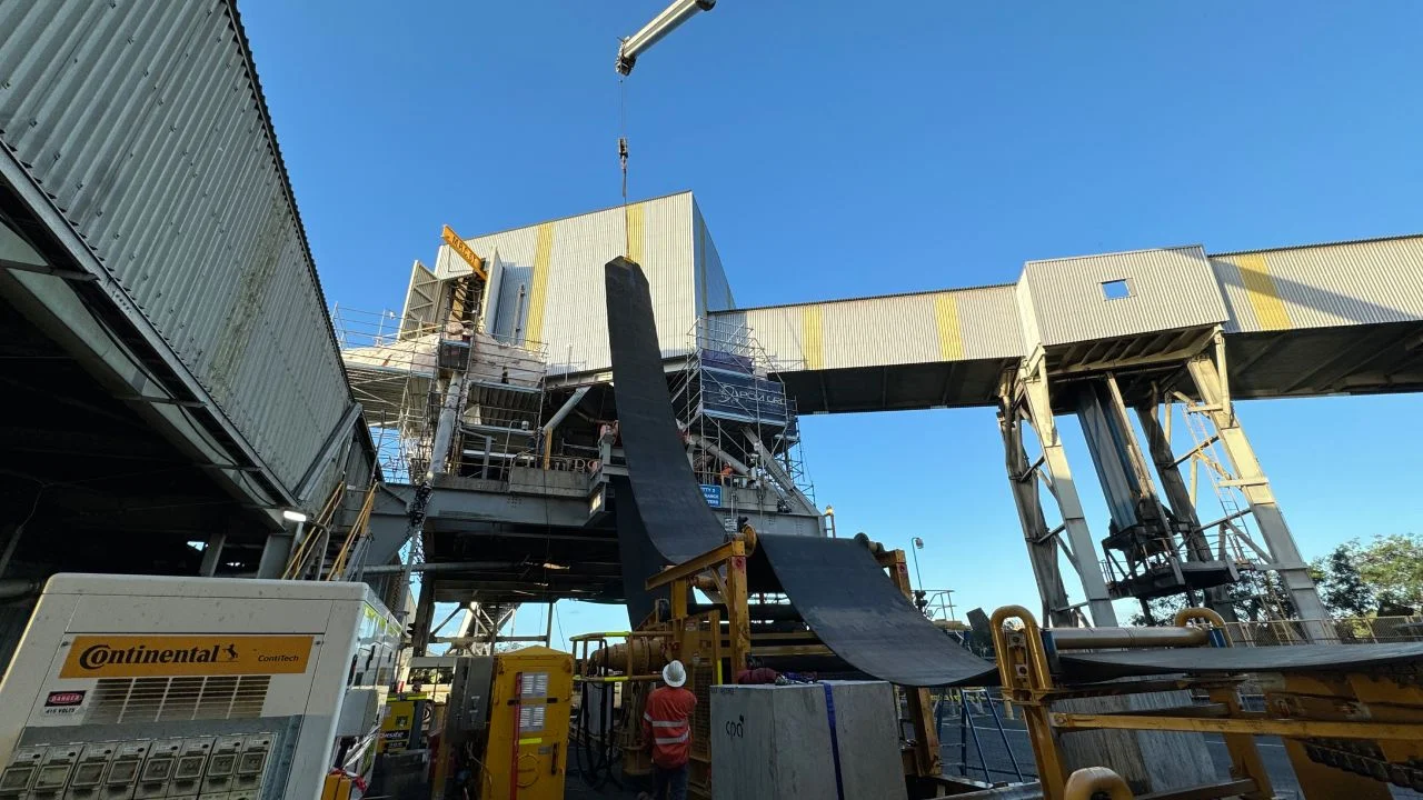 Workers and machinery install a large black conveyor belt at an industrial site with scaffolding, metal structures, and a crane under a clear blue sky. Safety equipment and supplies are visible in the area.