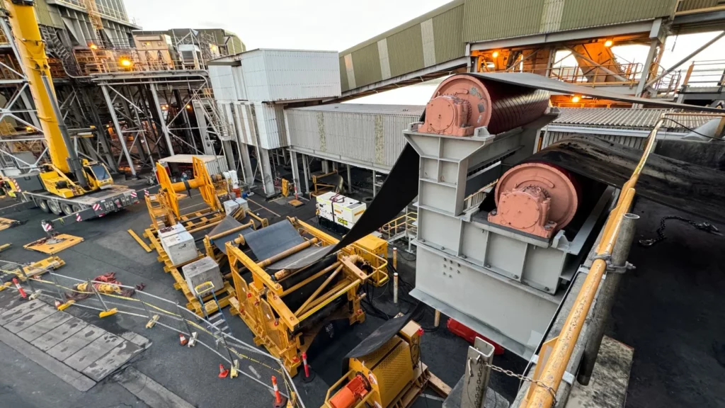 A large industrial facility with conveyor belts, machinery, and equipment. There are yellow barriers, construction vehicles, and structures with metal beams and corrugated panels under bright lights.