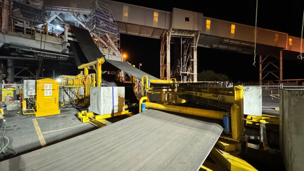 A nighttime industrial scene shows a large conveyor belt system transporting materials, with yellow machinery and structures illuminated by artificial lights under a covered area.