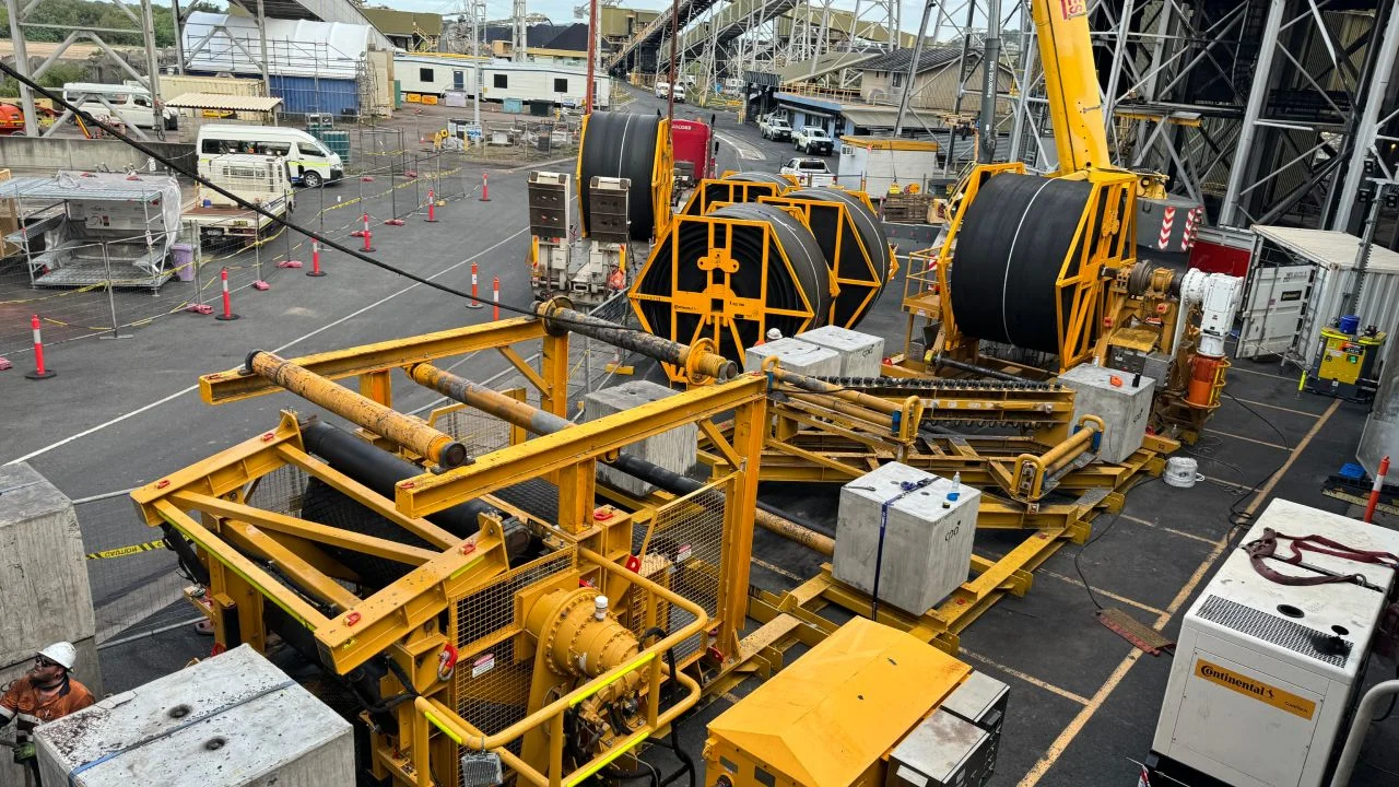 Large yellow industrial machinery with cables and spools is set up in a fenced-off area outdoors. Traffic cones, vehicles, and buildings are visible in the background. One worker in a helmet sits near the equipment.